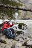Person in red jacket using Katadyn BeFree Gravity 3L water filter hanging from tree branch by river outdoors