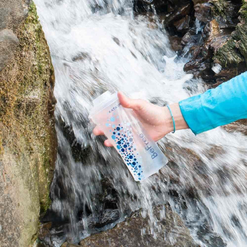 Hand holding Katadyn BeFree 0.6L water bottle filter under flowing stream water for instant filtration.