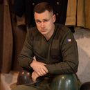 Man wearing original olive drab Czech army wool sweater with V-neck and flag patch on sleeve, sitting with helmets.